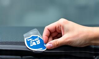 Eine Hand, die eine aufgeklebte Autobahn-Vignette korrekt an der oberen linken Ecke einer Windschutzscheibe positioniert.